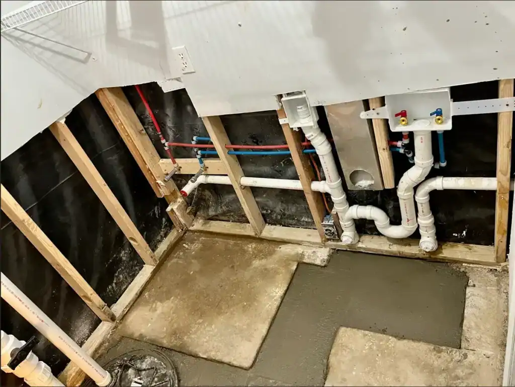 Exposed wall framing and plumbing in a partially finished bathroom, with white PVC pipes and concrete patches on the floor.