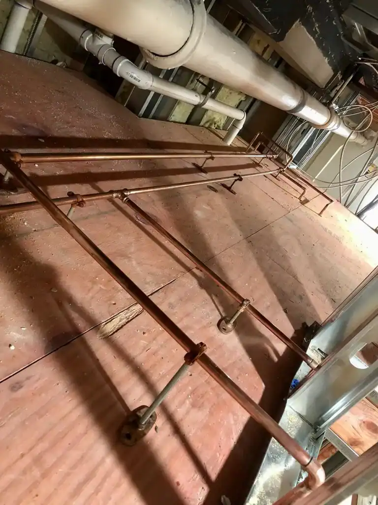 Rusty metal railing with crossbars on a pinkish metal floor, with exposed pipes overhead in an industrial setting.