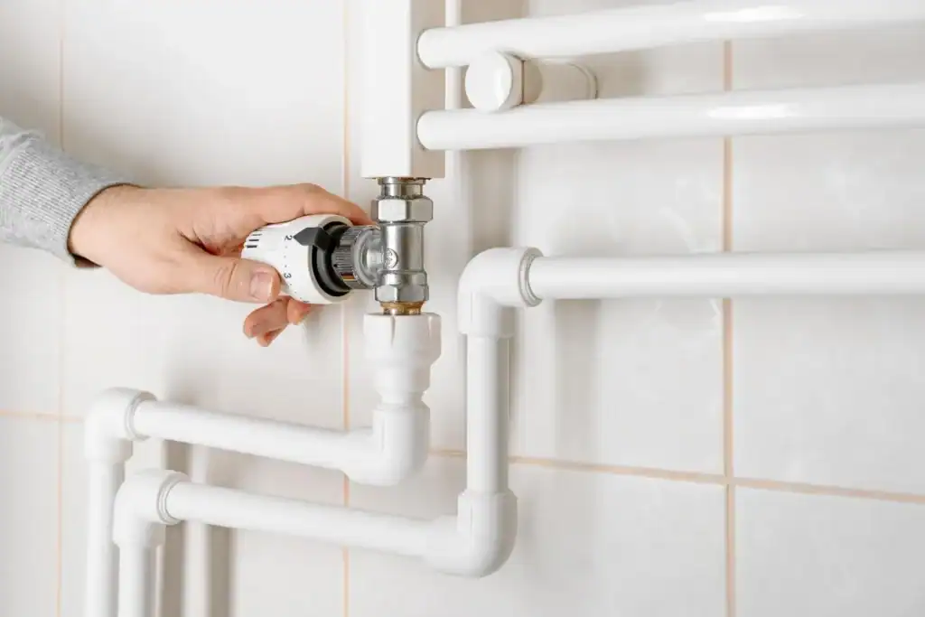 A hand turning a valve on white plumbing pipes mounted on a tiled wall.