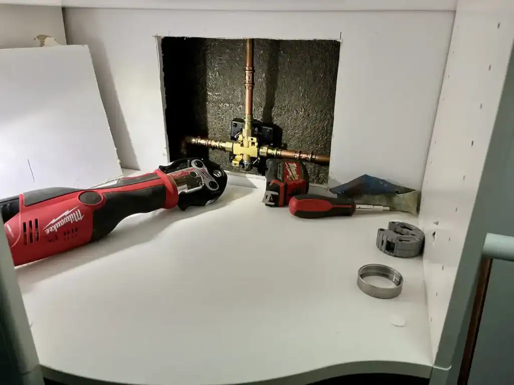 Open wall cavity with copper pipes and a red drill, with assorted tools and hardware on a white curved work surface.