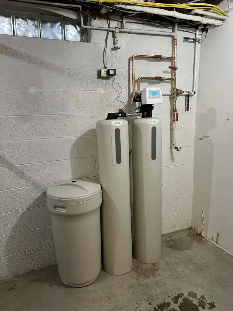 Basement utility area with two tall water filtration tanks, exposed piping and a control panel, beside a trash can.