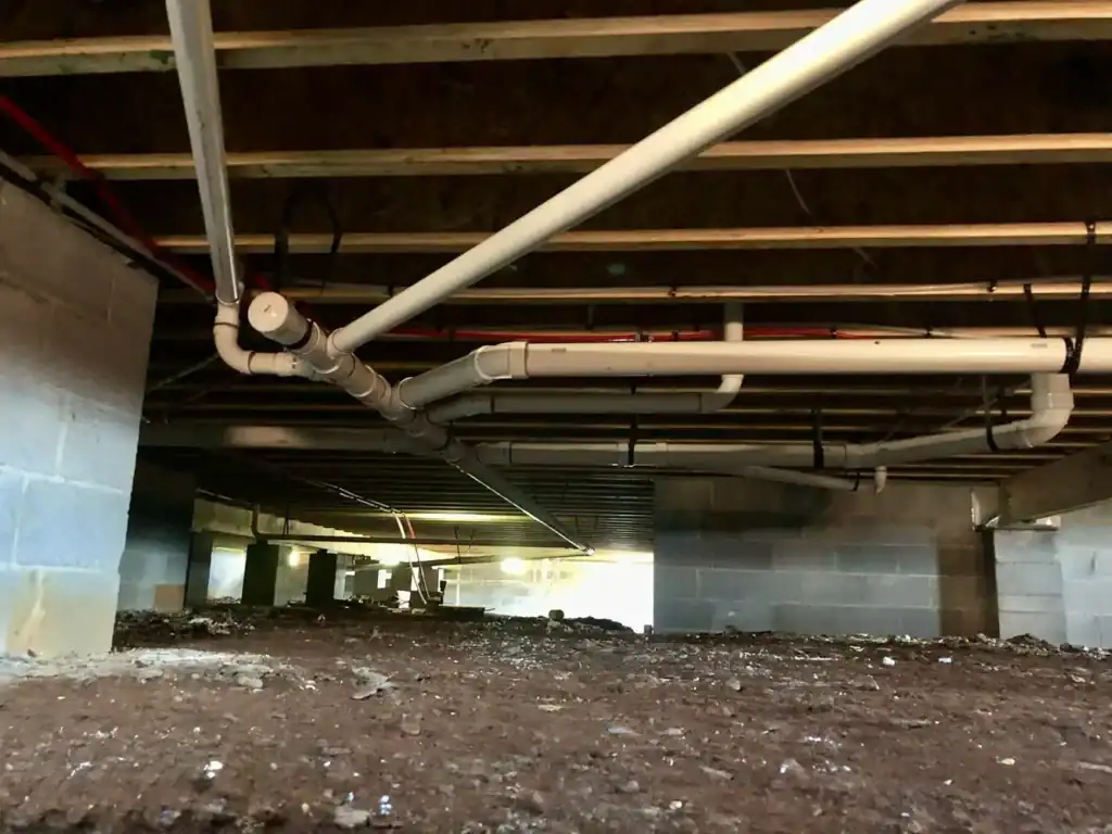 Crawl space under a building with exposed pipes and overhead beams, viewed from a dirt floor toward a distant light.