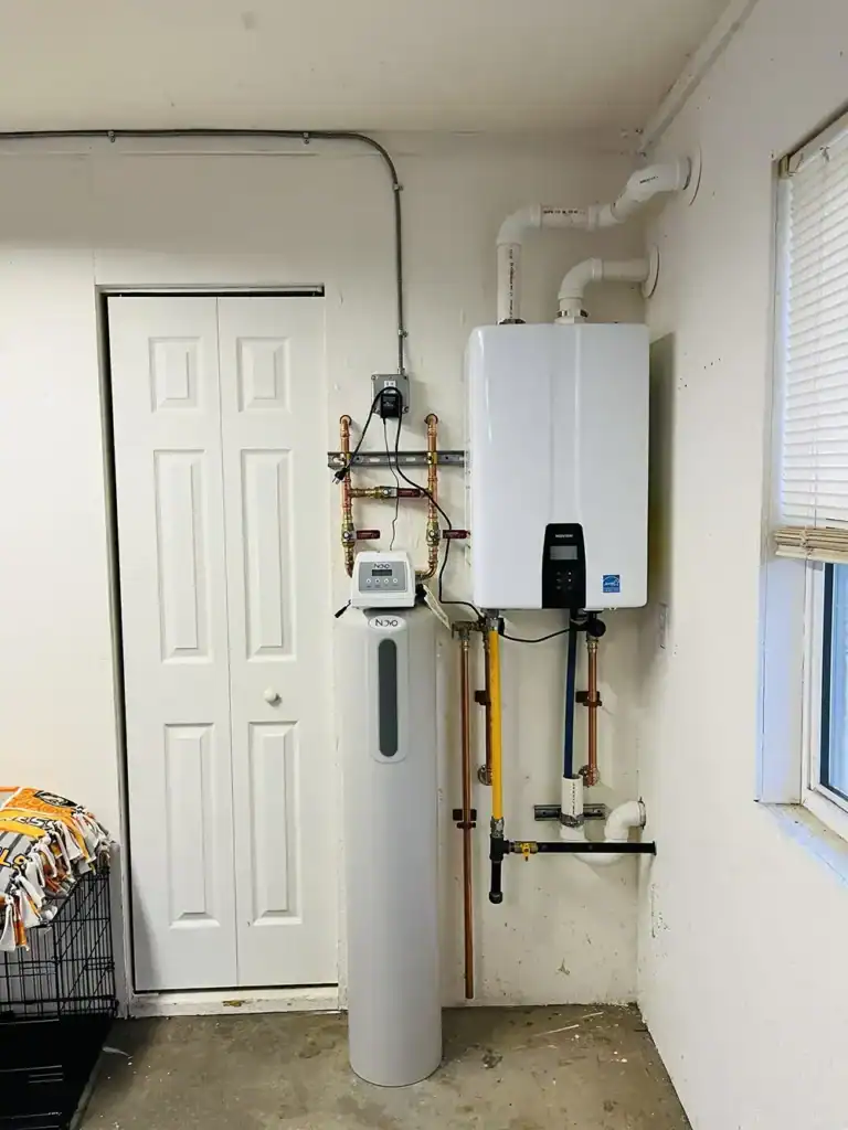 A home utility room featuring a tall gray vertical tank, copper piping, and a wall-mounted boiler beside a white door and a window.